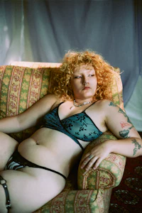 a white woman in black lingerie and blonde curly hair lying on a table. the table is covered a red silk table cloth.